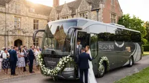 A wide landscape photograph of a modern, multi-axle dark grey Readybus corporate hospitality coach parked on a driveway in front of a grand historic English manor house. The front of the bus is elegantly draped in a white rose garland, with the bride, groom, chauffeur, and wedding guests posing together beside it during sunset.
