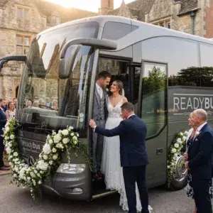 A newlywed bride and groom standing in the doorway of a dark grey luxury Readybus coach, looking lovingly at each other, while a chauffeur stands outside holding the door open against the backdrop of an old stone manor.
