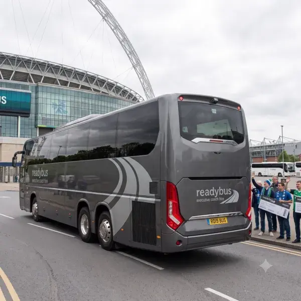 A rear three-quarter view of a sleek, charcoal-grey executive touring coach on a city street, with a massive curved stadium structure visible in the background under an overcast sky.
