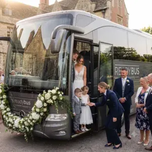 A professional female coach driver in a smart navy suit bending down to assist a young page boy and flower girl stepping off a grey luxury Readybus wedding coach decorated with white floral wreaths.