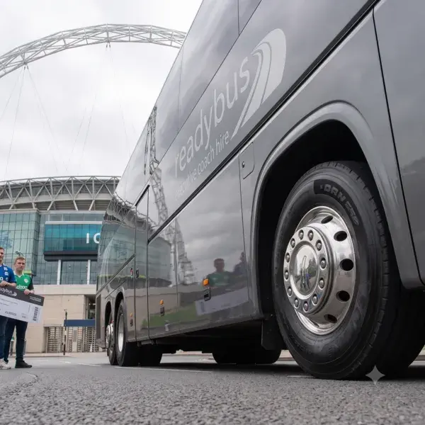 A low-angle, close-up shot focusing on the front wheel and polished chrome rim of a grey executive coach, showing the reflective side panel of the bus with a partial "readybus" logo, set against a stadium backdrop.