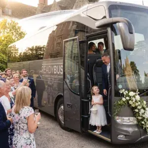 A close-up shot focusing on a young flower girl holding a basket on the steps of a grey Readybus wedding coach, with the smiling bride and groom standing just behind her inside the coach door as guests look on.