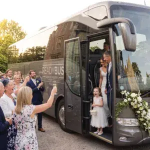 A joyous little flower girl in a white dress standing on the steps of an open door of a grey luxury Readybus coach, looking out excitedly at cheering wedding guests raising their champagne glasses.