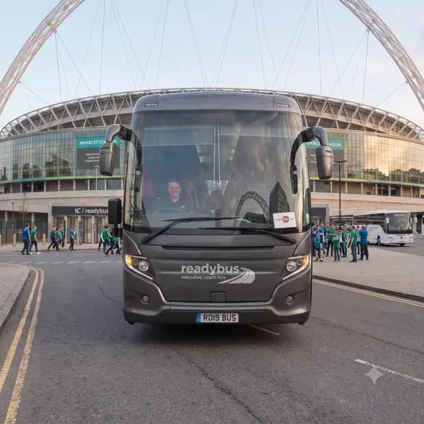 A front-facing view of a dark grey luxury coach driving down an open road toward the camera, with a large modern stadium and its distinctive white arch framing the background.