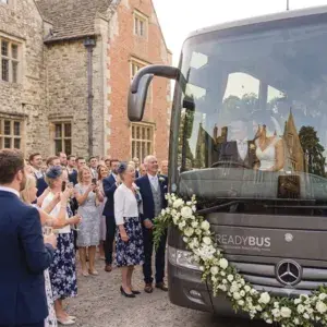 A frontal view of a dark grey luxury Mercedes coach decorated with a large garland of white roses. The bride and groom are visible sitting inside, with a clear reflection of a historic brick and stone manor house mirrored on the front windscreen.