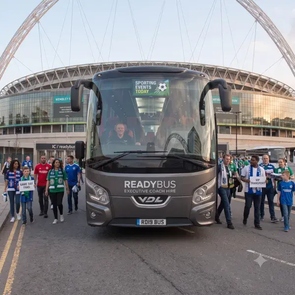 A front view of a grey executive coach on a stadium approach road, flanked by crowds of smiling sports fans walking alongside it wearing matching blue and green jerseys.