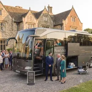 A side-angle view of a grey modern Readybus coach parked at a stately home wedding venue. The luggage bay is open showing wedding hampers, while well-dressed wedding guests talk amongst themselves on a gravel driveway during golden hour.